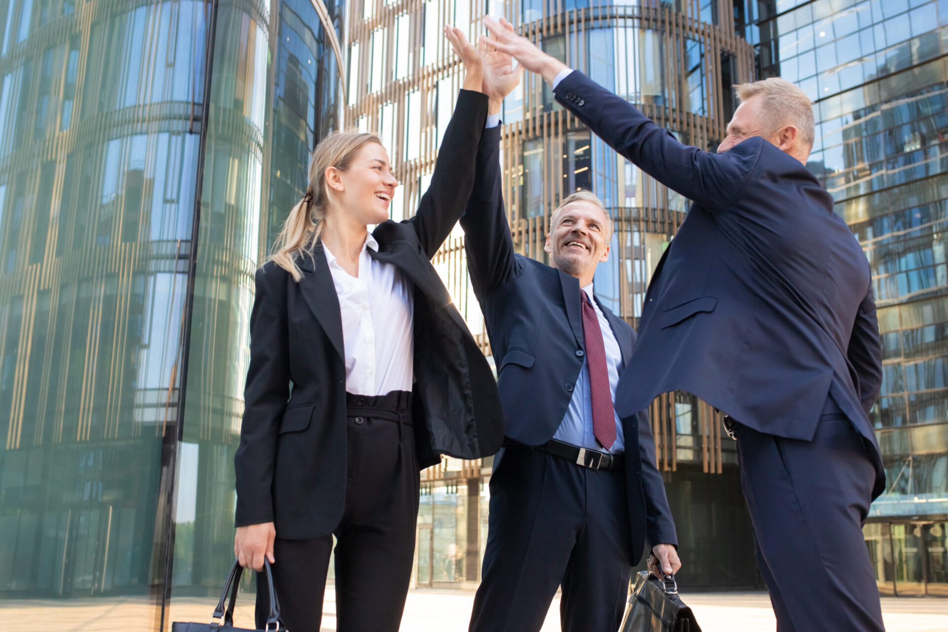 Three cheerful businesspeople giving five and smiling. Confident happy colleagues celebrating successful deal together, standing outdoors and rising hand up. Teamwork and partnership concept